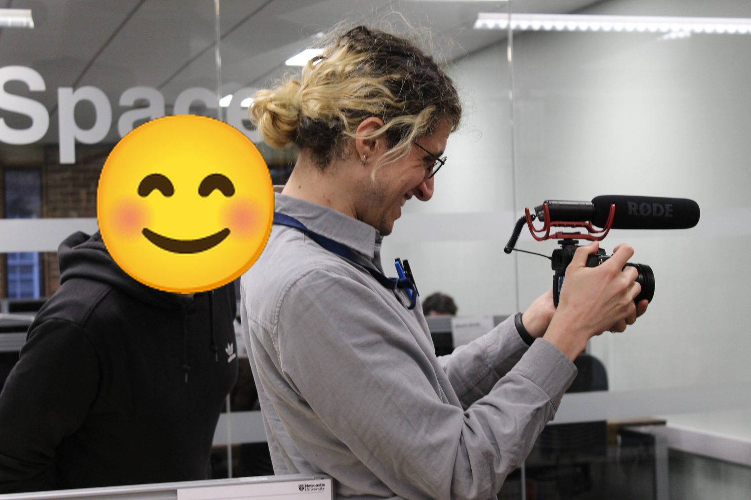 João Lazaro filming with a camera and shotgun microphone in a university space, a colleague watching over his shoulder