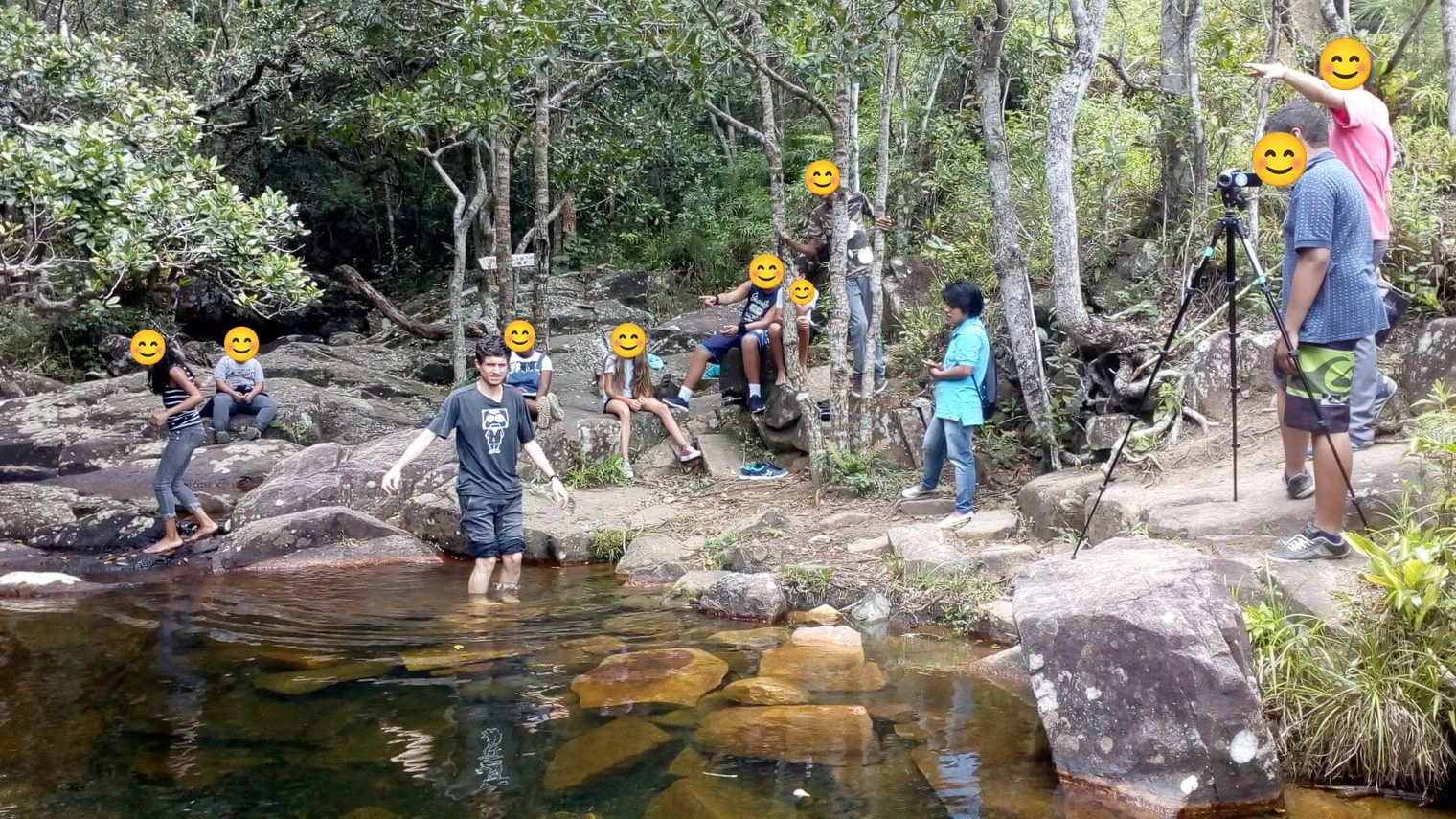 Educom.cine students and teachers filming A Maldição da Cachoeira by a river and waterfall, a camera on a tripod on the right; students' faces covered