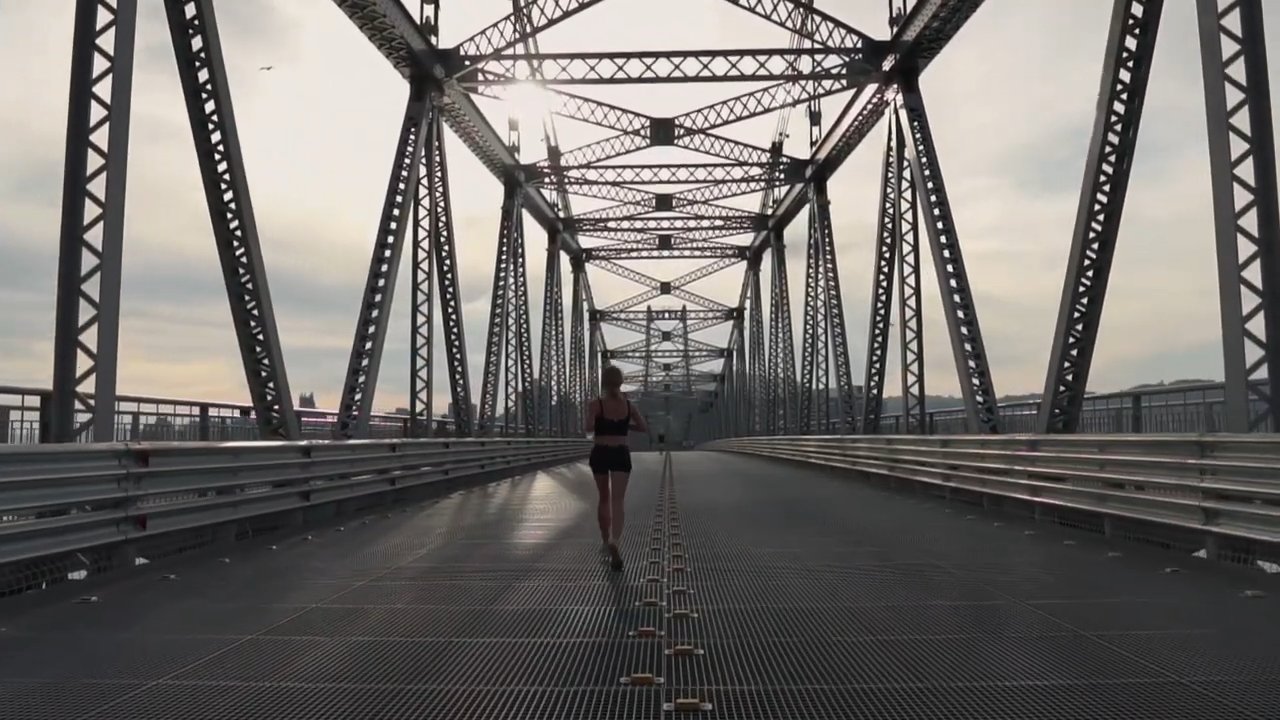 Still from Em Obras: a runner, seen from behind, alone on the restored deck of the Hercílio Luz Bridge, the steel trusses lined up to the horizon