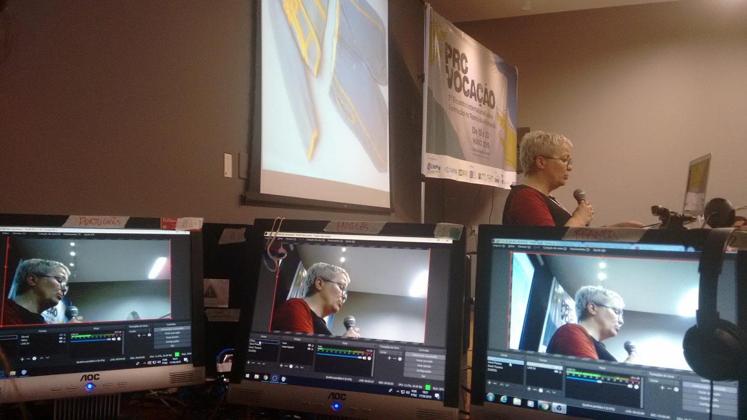 Three monitors showing the same speaker on the three language streams, with the speaker at the lectern and the PRO-VOCAÇÃO banner behind
